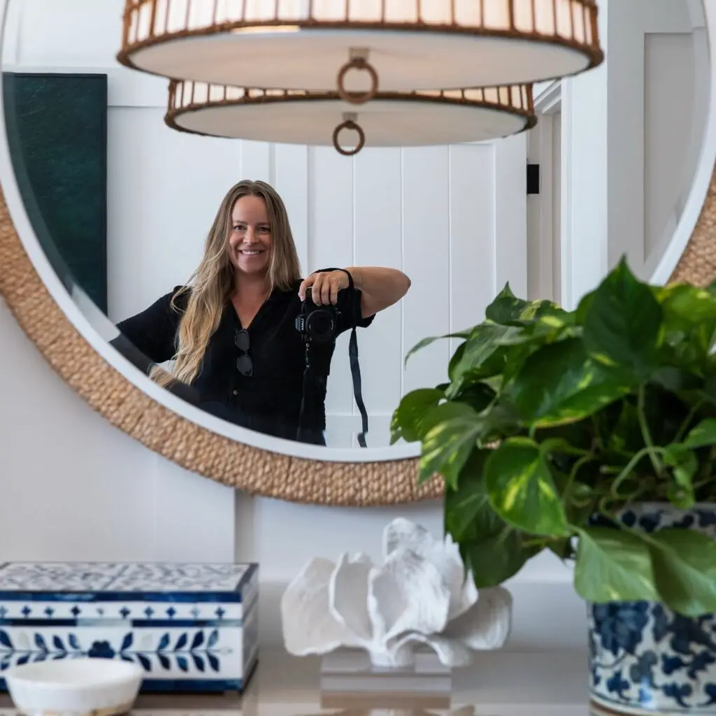 Megan Meek, interior photographer, reflected in a round woven mirror holding her camera in a styled coastal entryway with blue and white decor and indoor plants
